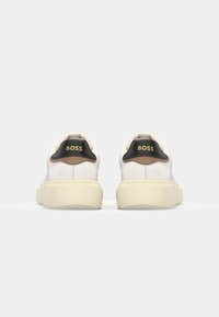 Witte leren sneakers met een gele zool, zwarte en bruine accenten bij de hiel, en het "BOSS"-logo in het geel op de achterkant.