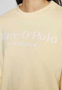 Sweat-shirt jaune en tissu doux, avec un col rond et un texte blanc indiquant "Marc O'Polo EST. À STOCKHOLM" sur le devant.
