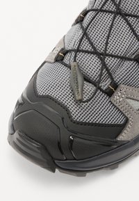 Szare buty sportowe z siateczkową cholewką, czarnymi gumowymi akcentami i teksturowaną podeszwą. Sznurówki są zabezpieczone za pomocą pętli dla większej wygody.