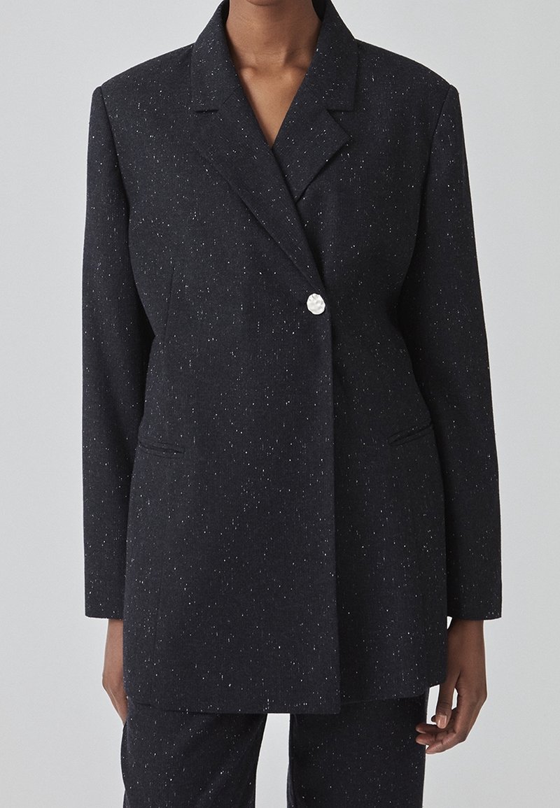 Blazer gris foncé tacheté avec un seul bouton blanc texturé, porté avec un pantalon assorti par une personne debout devant un fond uni.