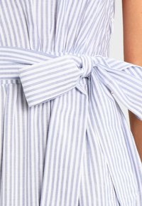 Un tejido de rayas azul claro y blanco presenta un cinturón de lazo, pliegues y una silueta fluida. La textura es suave y ligera, ideal para el clima cálido.
