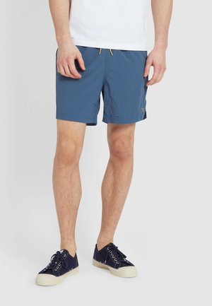 Blaue Badeshorts aus leichtem Material mit elastischem Bund und gelbem Zugband. Kombiniert mit dunkelblauen Canvas-Sneakern.