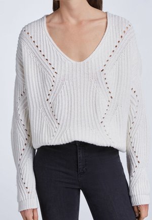 Frau trägt übergroßen weißen Strickpullover mit V-Ausschnitt und diamantförmigen, offen gestrickten Details, kombiniert mit schwarzen, eng anliegenden Jeans.