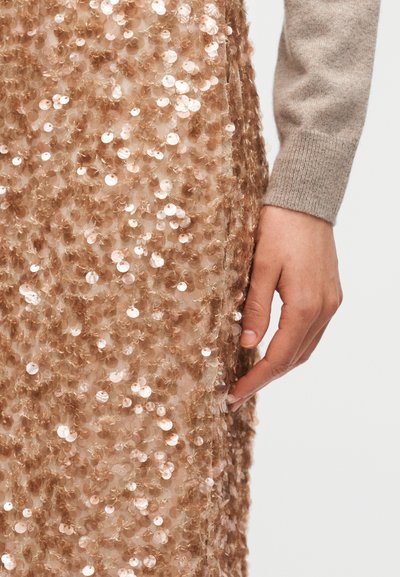 Φόρεμα με παγιέτες σε ανοιχτό καφέ απόχρωση, με πυκνό στρώμα από γυαλιστερές, στρογγυλές παγιέτες. Μια χέρι τοποθετείται κατά μήκος της άκρης του υφάσματος.