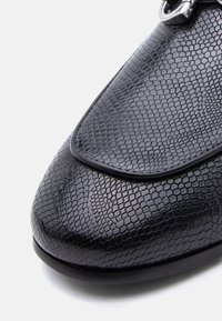 Μαύρο δερμάτινο παπούτσι με υφή που μοιάζει με ερπετό, με γυαλιστερή λεπτομέρεια στη μύτη και στρογγυλεμένο σχήμα.