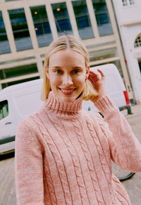 Light pink cable-knit turtleneck sweater with ribbed cuffs, featuring textured patterns. Worn with a backdrop of urban architecture.