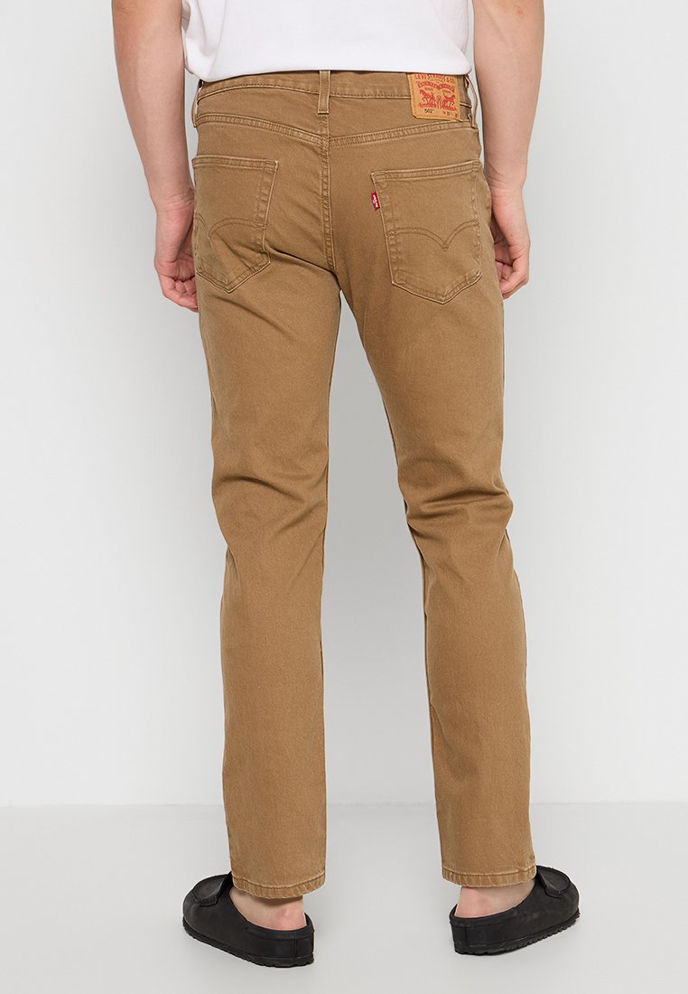 Uomo che indossa jeans Levi's beige e scarpe slip-on nere, in piedi contro un muro bianco, mostrato dalla vita in giù.