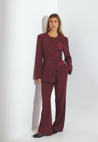 Burgundijas krāsas piegrieztais uzvalks ar dubulto jaku, kas satur se�šus pogas un taisnās kājas bikses, izgatavots no gludas auduma. Spīdīgas kurpes.