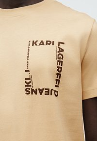Μπεζ μπλουζάκι με καφέ κείμενο "KARI LAGERFELD JEANS" διατεταγμένο σε τετράγωνο στο στήθος.