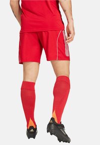 Rote Sportshorts mit weißen Seitenstreifen, kombiniert mit knielangen roten Socken mit orangefarbenen Akzenten und schwarzen Schuhen mit gelben Details.