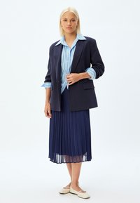 Marineblazer over een lichtblauw gestreept shirt, gecombineerd met een geplooide marineblauwe rok en flat creamkleurige schoenen, die een op maat gemaakt ontwerp en texturen tonen.