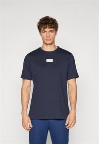 Camiseta de algodón azul marino de manga corta, con escote redondo y un parche de logo verde claro en el pecho. Textura suave, corte casual.