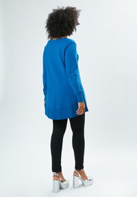 Langärmliger, Rundhals-Pullover in leuchtendem Blau, aus weichem Material; kombiniert mit schwarzen Leggings und silbernen Plateausandalen.