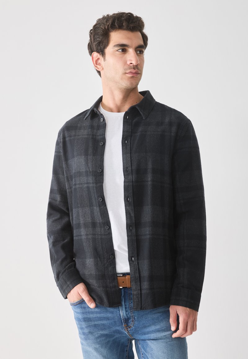 Knoop-up zwart en grijs geblokte flanellen shirt. Heeft een kraag, lange mouwen en een losse pasvorm, gecombineerd met lichtblauwe jeans en een bruine riem.