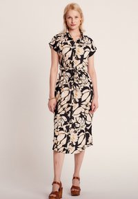 Robe à motifs floraux, à manches courtes, en noir, crème et or, avec un design à boutons et une taille ceinturée. Sandales à talons en bois.