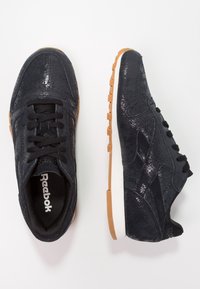 Svart Reebok-sneakers med en strukturerad faux ormskinnsövre, svarta snören och en sula av gummigummi. Vit mellansula erbjuder kontrast.