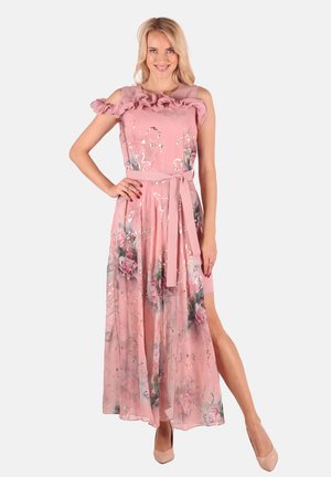 Lächelnde Frau in einem rosa geblümten Maxikleid mit Rüschen am Ausschnitt, seitlichem Schlitz und passendem Gürtel, trägt nudefarbene High Heels.