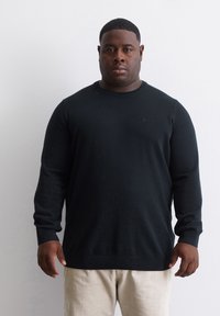 Schwarzer Strickpullover mit langen Ärmeln und ribbeligem Rundhalsausschnitt, versehen mit einem dezenten Logo. Kombiniert mit hellen Hosen.