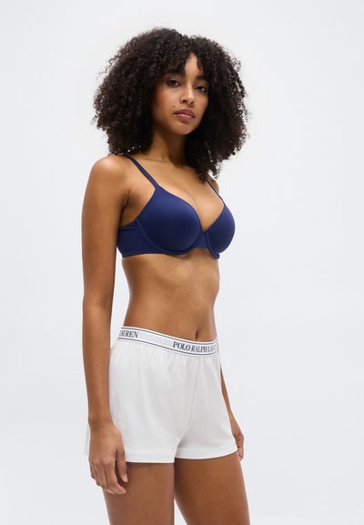 Γυναίκα με σγουρά μαλλιά που φοράει σουτιέν σε ναυτικό μπλε και άσπρα σορτς Polo Ralph Lauren, στέκεται μπροστά σε απλό φωτεινό φόντο.