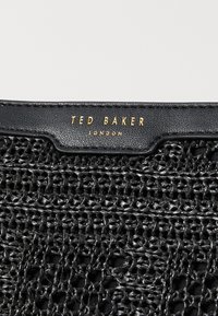 Crna tkanih torba s teksturiranom površinom, kožnim detaljima i zlatnim "Ted Baker London" logotipom na vrhu.