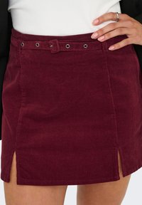 Burgundi väri corduroy mini seelik, millel on vööga talje, ees olevad lõhikuid ja nööpide detailid vöökoha juures, koos vertikaalse ribakudumise tekstuuriga.