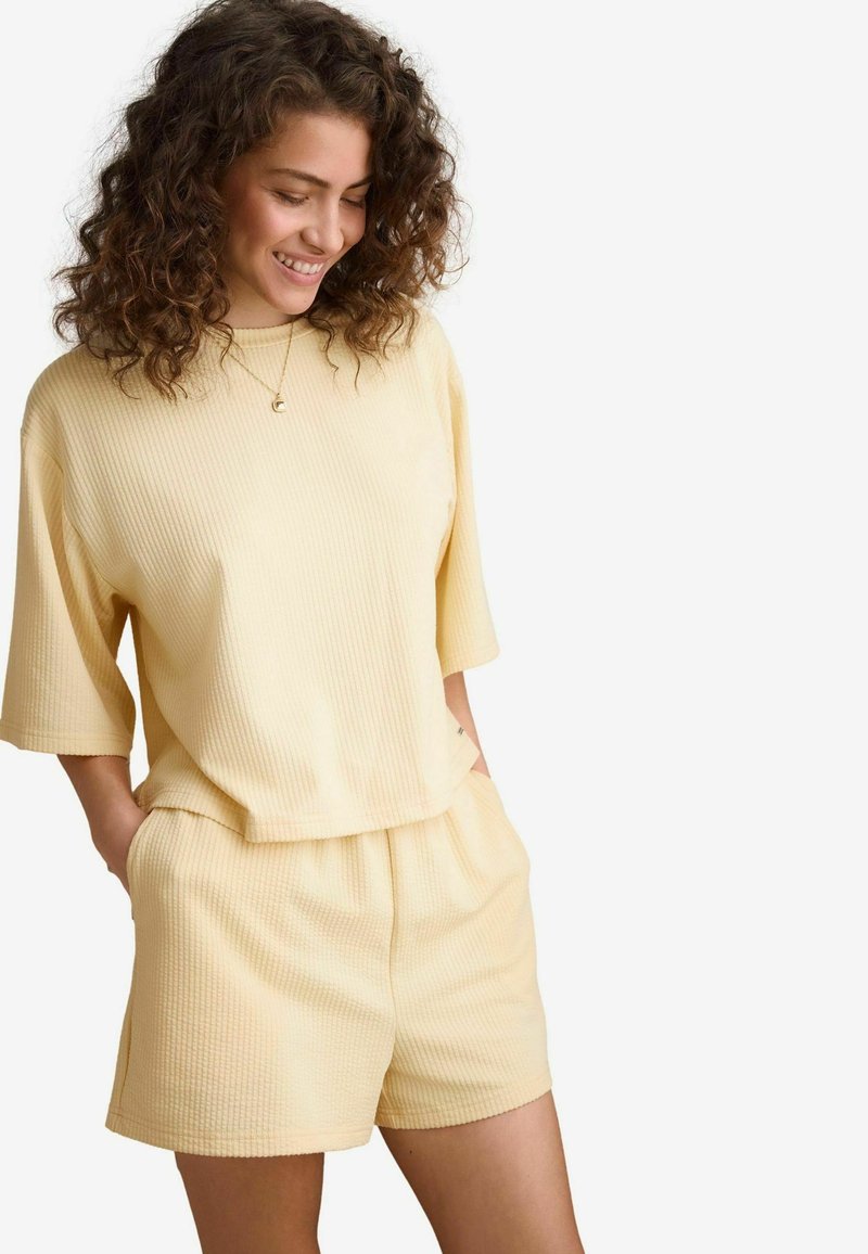Kollane ribblikuga lühikeste varrukatega tops, mis on kombineeritud sobivate lüh pükstega. Kangas on pehme tekstuuriga, sisaldades elastset vöökohta ja küljetaskuid.