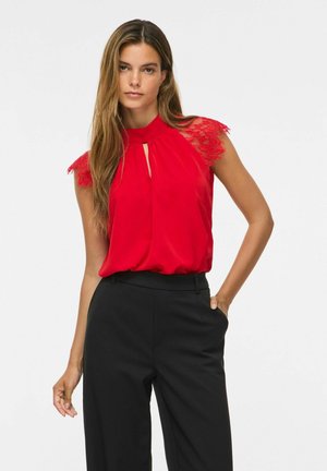 Femme aux longs cheveux bruns portant un chemisier rouge sans manches avec des épaules en dentelle et un pantalon noir taille haute, debout avec une main dans la poche.