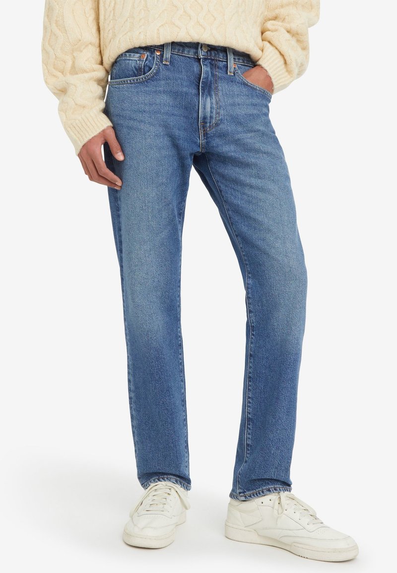 Jean en denim bleu clair avec une coupe droite, cinq poches et une texture légèrement délavée. Associé à des baskets blanches pour le contraste.
