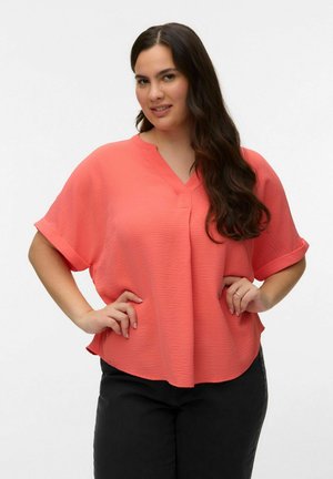 Žena s dugom tamnom kosom, odjevena u koraljnu bluzu kratkih rukava s teksturom i crne hlače, stoji s rukama na bokovima, blago se smiješeći.