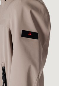 Giacca beige esposta su una spalla, presenta una toppa nera con accenti rossi, texture liscia, dettagli con zip, design moderno.