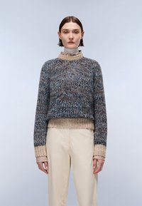 Sweter dzianinowy o teksturowanym wzorze w odcieniach niebieskiego, brązowego i kremowego, z wysokim kołnierzem i ściągaczami, w połączeniu z jasnymi spodniami.