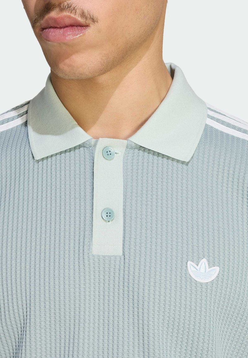 Mann trägt ein hellgraues, strukturiertes Poloshirt mit zwei Knöpfen und weißem Adidas-Trefoil-Logo auf der Brust.