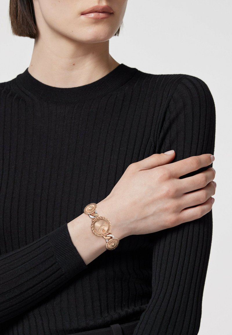 Gold-tone bracelet with circular and oval links, featuring textured details. Worn on a wrist against a black ribbed sweater background.