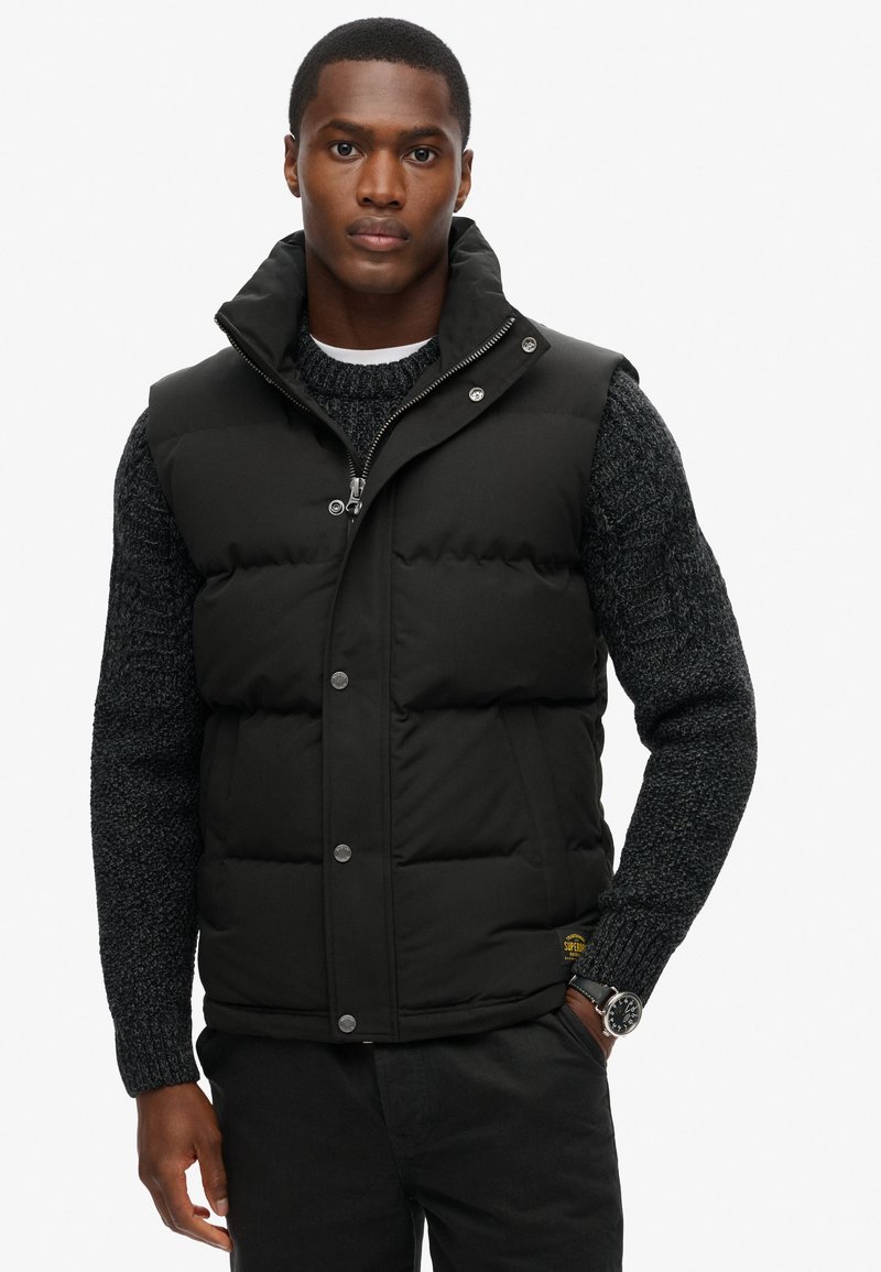 Hombre vestido con un chaleco acolchado negro sobre un suéter de punto gris oscuro, pantalones negros y un reloj de pulsera, de pie con una mano en el bolsillo.