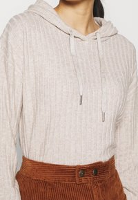 Sudadera beige de canalé con capucha y cordones, combinada con pantalones de pana color óxido que cuentan con dos botones en la cintura.