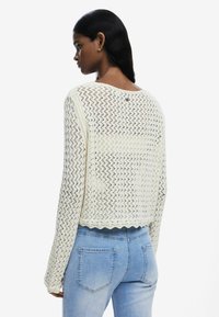 Strickpullover in Creme mit wavy Muster, langen Ärmeln und gewelltem Saum, getragen über hellblauen Jeans. Hat einen runden Ausschnitt.