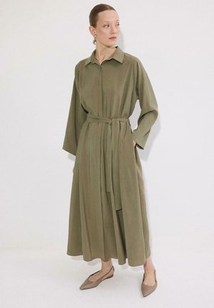 Γυναίκα ντυμένη με μακρυμάνικο ελαιί φόρεμα με ζώνη και γιακά, όρθια με τα χέρια στις τσέπες, μπεζ μυτερά φλατ παπούτσια και χρυσούς κρίκους σκουλαρίκια.