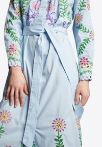 Abito azzurro chiaro con ricami floreali in rosa, giallo, viola e verde, con vita legata e polsini a scallop. Materiale in cotone, maniche lunghe.