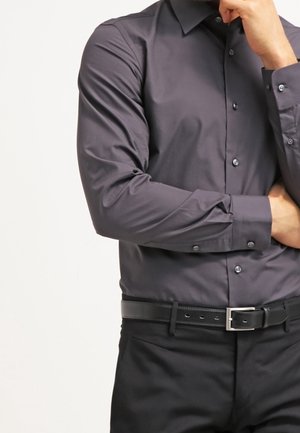 Mann trägt ein eng anliegendes dunkelgraues Hemd mit Knöpfen und schwarze Hose mit schwarzem Ledergürtel, Hand ruht nahe am Kinn, unter den Schultern beschnitten.