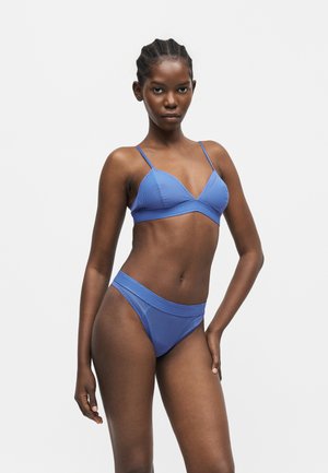 Žena v modrých rebrovaných trojuholníkových bikinách s tenkými ramienkami pózuje na jednoduchom svetlom pozadí.
