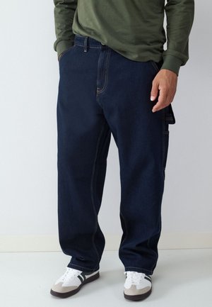Person wearing dark blue loose-fitting jeans, green long-sleeve shirt, and white sneakers with black stripes, standing with one hand in pocket.
