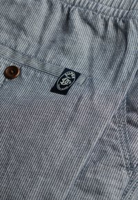 Grijs en witte gestreepte shorts van zacht materiaal, met een bruine knop en een marineblauwe label met witte tekst. Inclusief een zijzak.