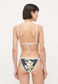 Rövid sötét hajú nő fehér pántos bikini felsőben és virágmintás bikini alsóban, háttal áll egy sima háttér előtt.