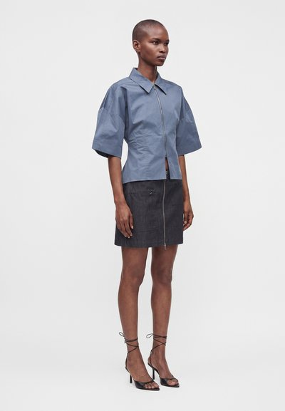 Femme debout portant une chemise bleue à manches courtes avec fermeture éclair, une jupe noire zippée et des sandales à talons hauts lacées noires sur un fond uni.
