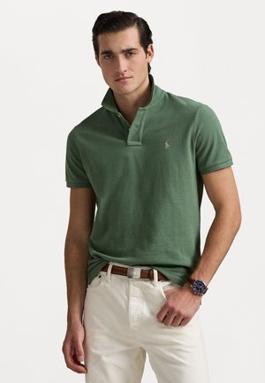 Groene polo met een gestructureerde stof, een drieknoopssluiting en een klein geborduurd logo. Gecombineerd met witte broeken en een bruine riem.