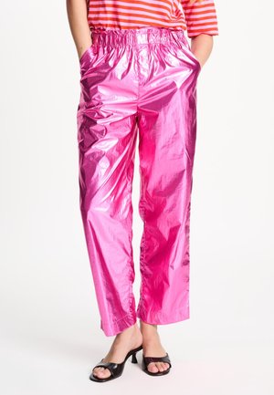 Persoon draagt glanzende metallic roze broek met elastische tailleband, handen in de zakken, gecombineerd met zwarte open-toe hakken en een rood gestreept topje.