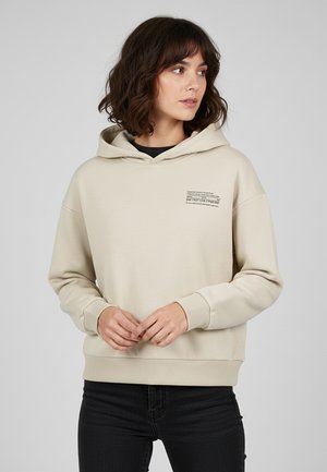 Beige hooded sweatshirt made of soft fabric. Features a front pocket, ribbed cuffs, and a graphic print on the chest. Model wears black jeans.