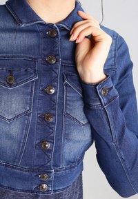 Denimjacka med en mörkblå gradient, silverfärgade knappar, dubbla bröstfickor, en avkortad design och sydda detaljer.