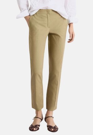 Donna che indossa pantaloni beige su misura alla caviglia, camicetta bianca con dettaglio in pizzo e tacchi marroni aperti con cinturini, in piedi con le mani parzialmente nelle tasche.