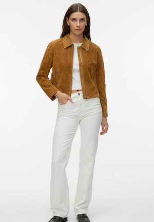 Femme portant une veste en daim marron zippée, un top court blanc, un pantalon blanc taille haute et des chaussures noires, debout avec une main dans la poche.